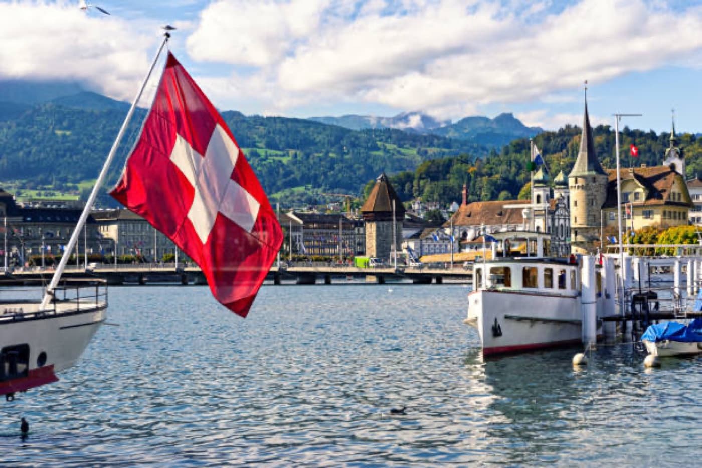 Swiss flag flying over Lake Lugano with the town and Alps behind
