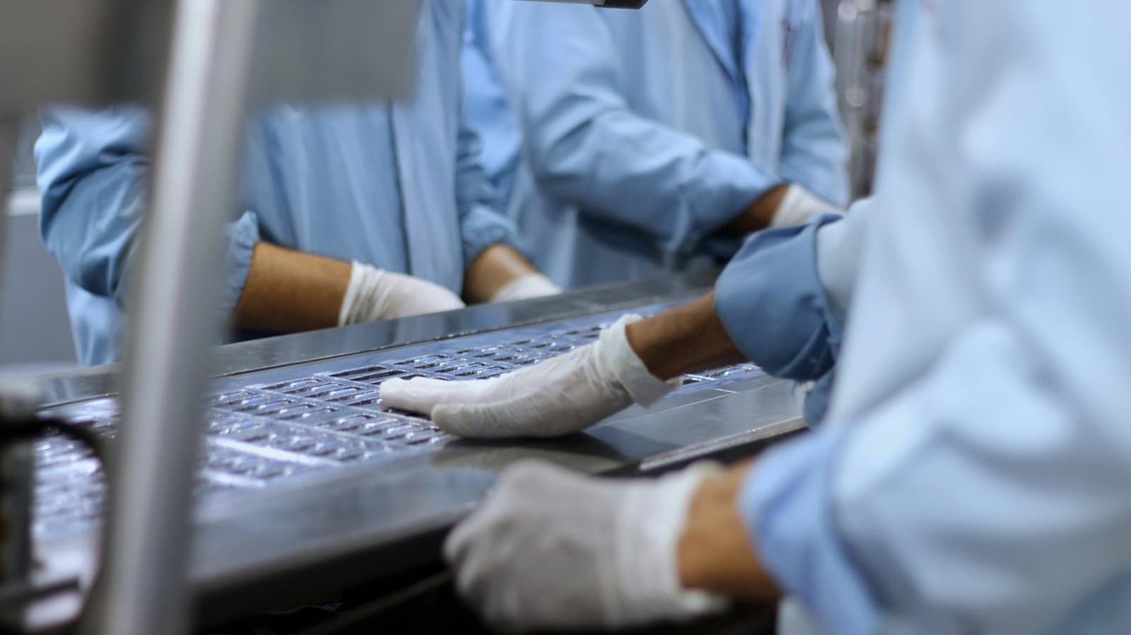 Workers in cleanroom smocks and gloves inspecting blister-packed tablets on a stainless pharmaceutical production line.